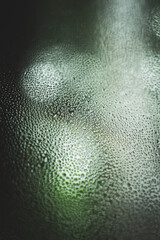 Close-up of water droplets on a fogged glass surface with soft green bokeh lighting in the background. The abstract texture and moisture pattern evoke calmness and a serene natural ambiance.