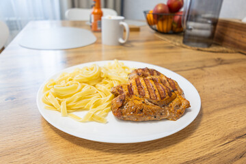 Grilled pork steak with pasta served on a white plate on wooden kitchen table.