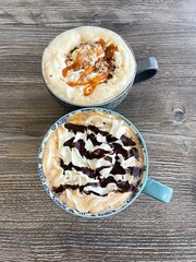 Two fancy gourmet coffees on a rustic wooden background. One coffee is a caramel macchiato and one is a cafe mocha drizzled with chocolate.