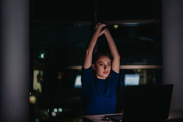 A young businesswoman stretches at her desk while working late into the night. Her focus and...
