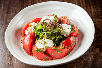 A fresh salad with ripe tomato wedges, creamy cheese, mixed greens, and a sprinkle of herbs and seeds, served in a white bowl on a rustic wooden table