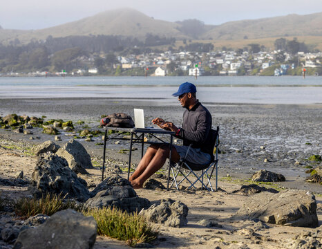 A man with a laptop working on a beach or bay at low tide.  Full-time RV nomad lifestyle, working in shorts from his home office.