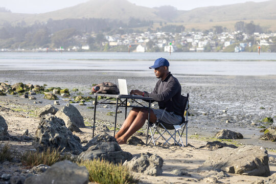 A man with a laptop working on a beach or bay at low tide.  Full-time RV nomad lifestyle, working in shorts from his home office.
