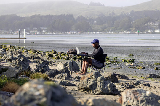 A man with a laptop working on a beach or bay at low tide.  Full-time RV nomad lifestyle, working in shorts from his home office.
