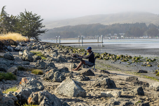 A man with a laptop working on a beach or bay at low tide.  Full-time RV nomad lifestyle, working in shorts from his home office.