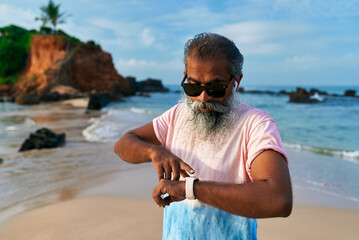 Senior ethnic man with grey beard on beach uses smart watch and earbuds for fitness. Elderly male enjoys tech for training, wellbeing. Old man in modern scene healthy lifestyle.