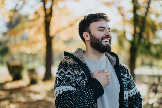A cheerful man in a warm, patterned sweater stands amidst autumn trees, laughing freely. The sunlit background and fallen leaves capture the essence of an enjoyable, relaxed outdoor moment.