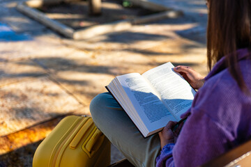 Young female traveler reading a book next to her suitcase in urban square.