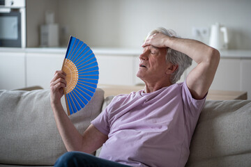 Elderly man feeling dizzy and unwell from heat, fanning himself at home without cooling system. Suffering from stuffy conditions, headache nausea high temperatures, senior vulnerability to heatwaves