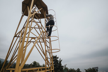 An individual ascends a symmetric yellow metal tower under an overcast sky, surrounded by trees on the ground, emphasizing adventure, risk, and outdoor exploration.