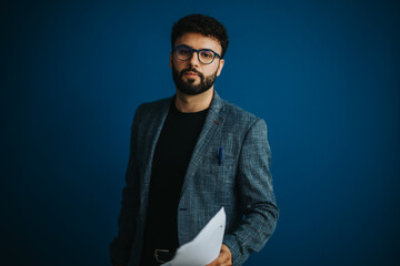 Portrait of a confident professor holding documents against a blue background. Captures...