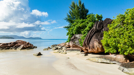 A peaceful beach with soft sand, large rocks, and vibrant green trees against a calm blue ocean. Praslin, Seychelles.