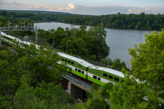 Aerial view of a train going through a lush green forest area by the lake in summer time at sunset hour. Commute, public transit, green transport, zero emission, travel, journey concept. 