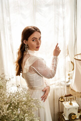 Elegant young woman in a vintage dress gazes through sheer curtains by soft sunlight