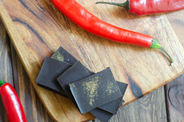 Pieces of extra dark chocolate with three red chili peppers on a wooden cutting board on the table. Trend food concept. Selective focus. Horizontal orientation. Top view