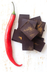 Pieces of extra dark chocolate with red chili peppers on a white wooden cutting board on the table. Trend food concept. Selective focus. Vertical orientation. Top view.