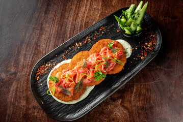A gourmet dish of crispy potato pancakes topped with smoked salmon, garnished with fresh herbs and green onions, served on a sleek black plate with a side of cucumber salad
