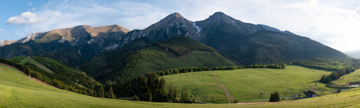 Panoramic view of High Tatras peaks including Havran and Ždiarska vidla above green meadows and forest near Ždiar, Slovakia, with ski lift structures visible in the landscape.