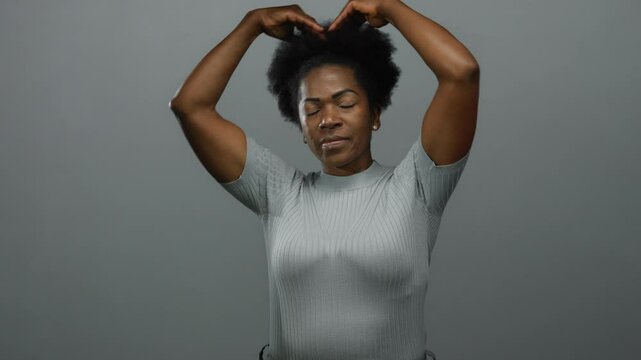Woman making heart gesture with hands against a grey background featuring a young african american expressing joy and love in a simple serene setting.