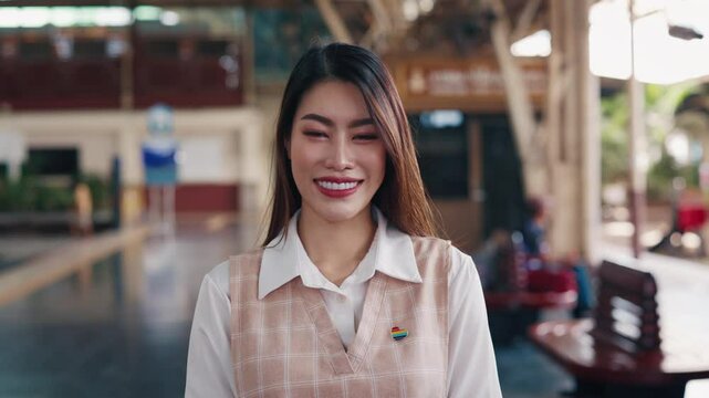 Portrait of positive Asian pride woman with rainbow heart pin on vest at train station. Pretty smiling lesbian shows proudness of LGBTQI community travelling by railroad transport