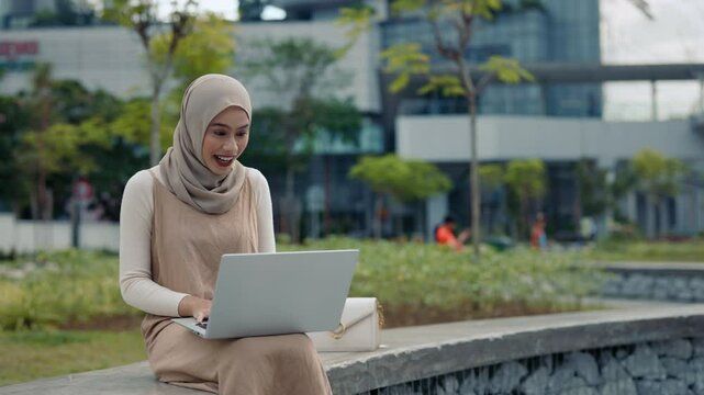 Portrait of joyful Malaysian woman with hijab joins online meeting via laptop sits in city park. Young muslin lady greets colleagues on video chat via notebook computer on street