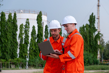 Workers in orange uniforms collaborating on a laptop at an industrial site surrounded by greenery...