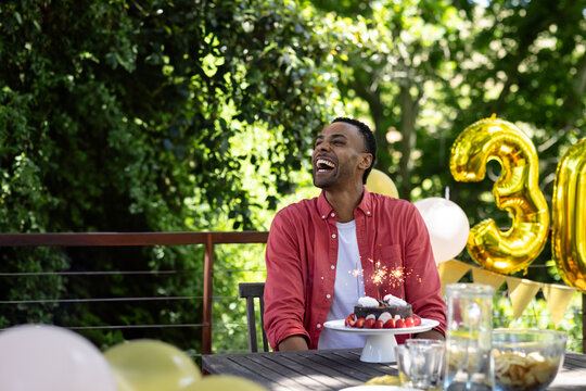 African American man sitting at patio table celebrating thirtieth birthday with cake and balloons - Powered by Adobe