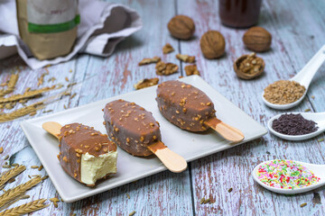 Bitten vanilla ice cream bar with chocolate and nut coating on ceramic plate. Rustic wooden background, surrounded by natural ingredients
