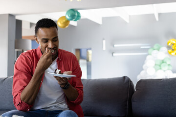 Mid adult African American man sitting on sofa eating cake under balloons at home, copy space
