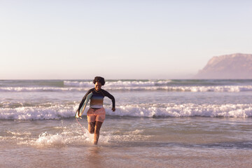 African American woman running from surf toward sandy shore holding surfboard wearing rash guard