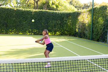 Hispanic girl wearing pink sportswear with racket focusing on yellow ball over net on tennis court