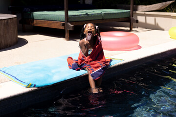 African American girl sitting wrapped in red towel at pool edge licking popsicle under sunlight