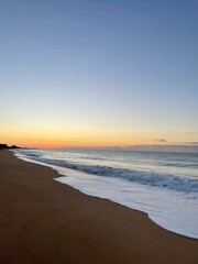 Sunset at the shore in Albufeira, Portugal. Gentle Atlantic waves roll in with white foam as the sky glows orange and yellow above the horizon. A peaceful coastal scene.