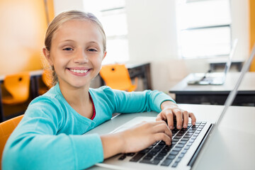 Student typing on silver laptop at bright classroom desk with orange chairs and daylight streaming