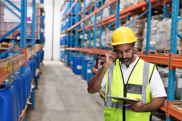 Man wearing yellow helmet and vest using clipboard speaking on phone in warehouse aisle, copy space