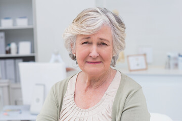 Senior woman sitting in clinic facing camera with computer monitor, binders and medical supplies