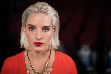 Woman gazing at camera posing in dark auditorium wearing red blouse and gold-and-pink necklace