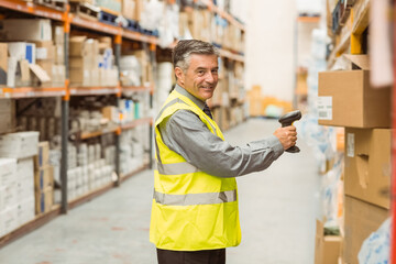 Middle-aged man wearing safety vest scanning boxes with handheld barcode scanner in warehouse aisle