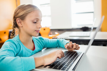 Child typing on laptop at desk in school classroom, with orange chairs and sunlight