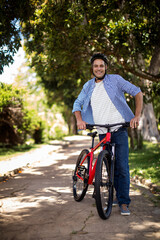 Obraz premium Man standing holding red mountain bicycle on sunlit tree-lined path wearing cycling helmet