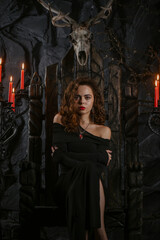 Fototapeta premium Gothic woman in black dress posing as a queen on anceint throne in candlelight on dark background.