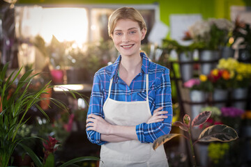 Florist wearing blue plaid shirt and beige apron standing arms crossed among plants in sunlit shop