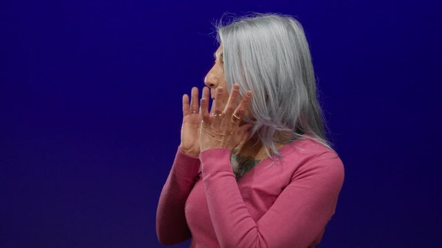 Senior grey-haired woman in a pink shirt looking surprised against isolated blue background, capturing candid emotional expression.