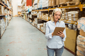 Woman standing in warehouse aisle scanning inventory on clipboard and wearing headset, copy space