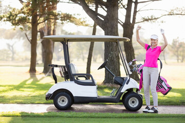 Woman in pink polo celebrating beside cart and clubs on golf course path, copy space