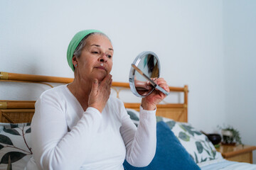 Elderly woman taking care of her face skin sitting on bed, applying moisturizing cream