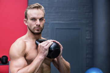 Shirtless muscular man lifting black kettlebell at chest height in gym area with dumbbell rack