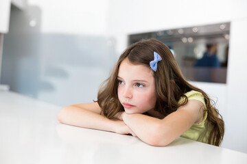 Obraz premium Female child leaning on glossy white countertop in kitchen wearing blue hair bow gazing at oven