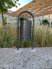 Cylindrical metal street urn, fixed between two arc-shaped posts, set on concrete surface, against background of tall dry grass, ahead of modern building with beige facades and canopy