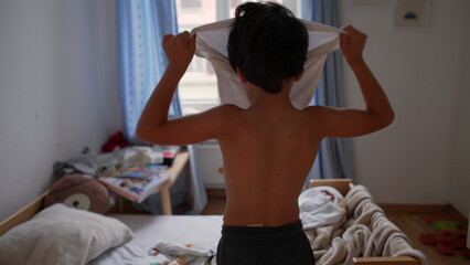 Young boy putting on his shirt in the bedroom, independent child dressing himself during morning...